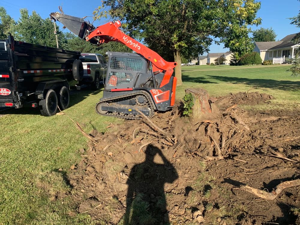 Stump grinding equipment in a yard, with uprooted tree remnants and a shadow in the foreground.