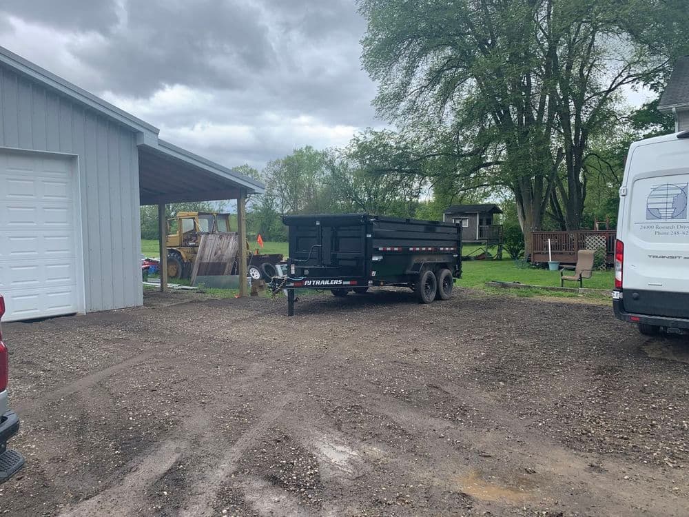 Dump trailer parked on gravel lot beside a grey shed and trees, with equipment nearby.