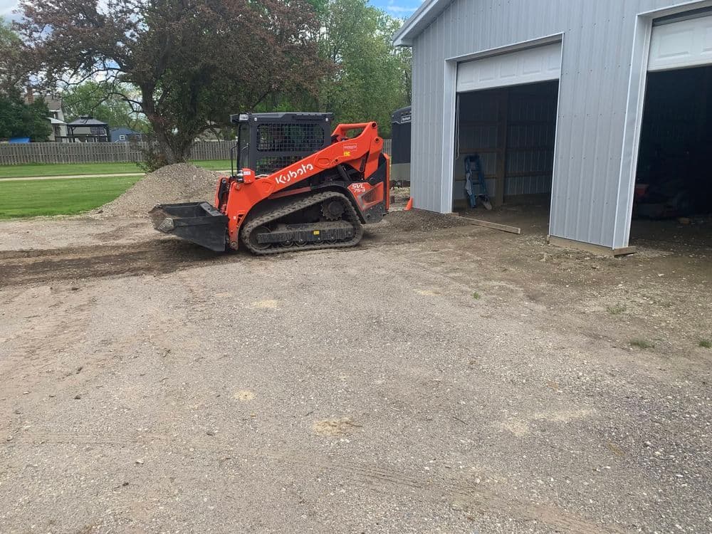 Orange compact track loader operating near a shed on a gravel surface.