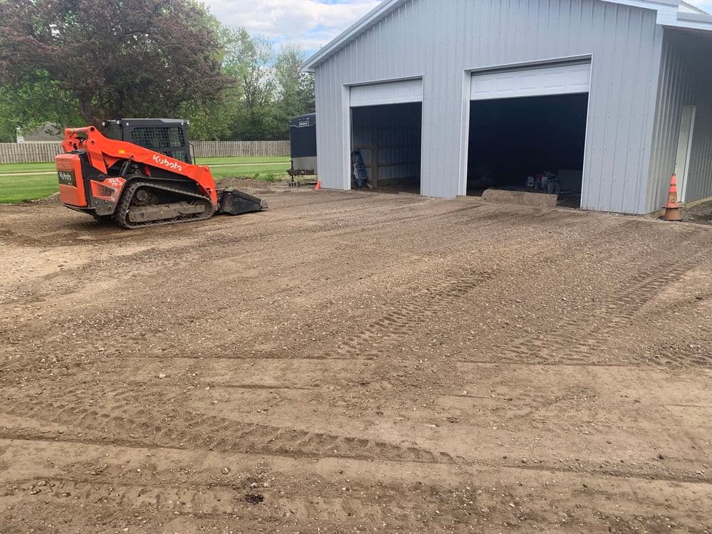 Bobcat skid steer on gravel driveway beside two gray storage garages. Freshly graded surface.