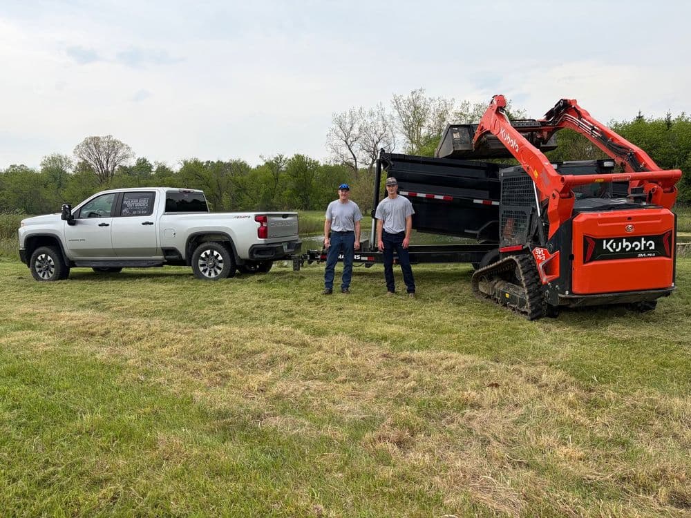 Two workers stand beside a Kubota mini-excavator and a silver pickup truck on a grassy field.